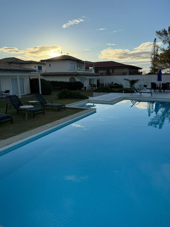 Piscina ampla ao entardecer, com espreguiçadeiras no gramado e casas de dois andares ao fundo.