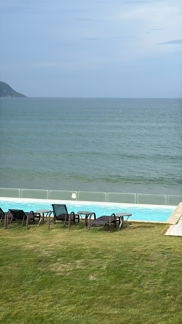 Piscina com fechamento de vidro de frente para o mar, com espreguiçadeiras sobre o gramado.