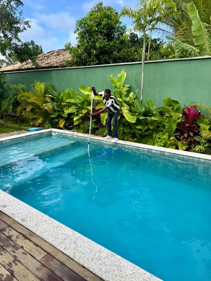 Profissional em pé na borda aspira o fundo de uma piscina de água azul, com jardim tropical e muro verde ao fundo.